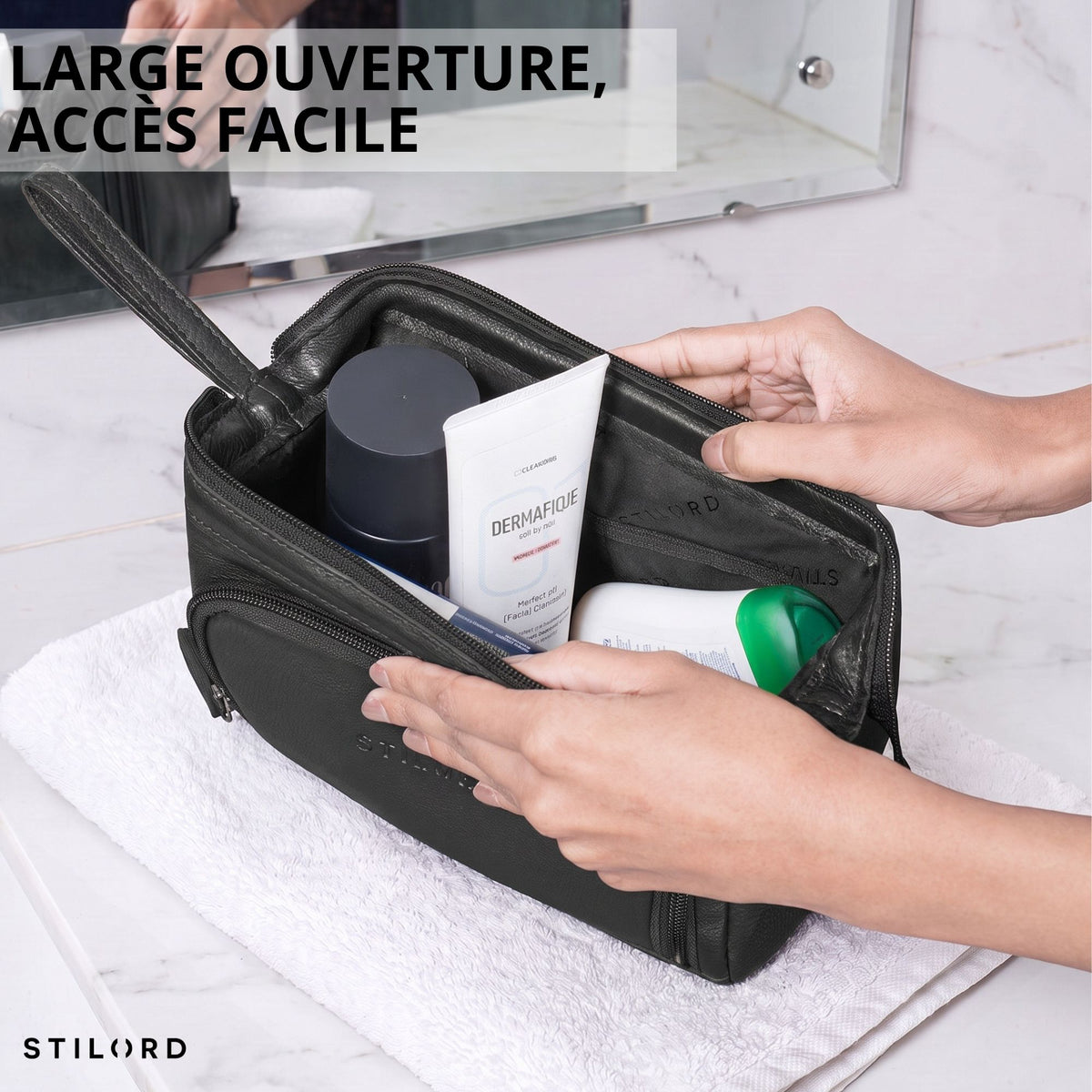 Trousse de toilette en cuir noir ouverte, vue intérieure avec produits, mains ouvrant la fermeture, logo STILORD embossé visible