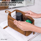 Trousse de toilette en cuir tan ouverte sur une serviette, mains écartent la fermeture, produits de soin visibles, marquage STILORD