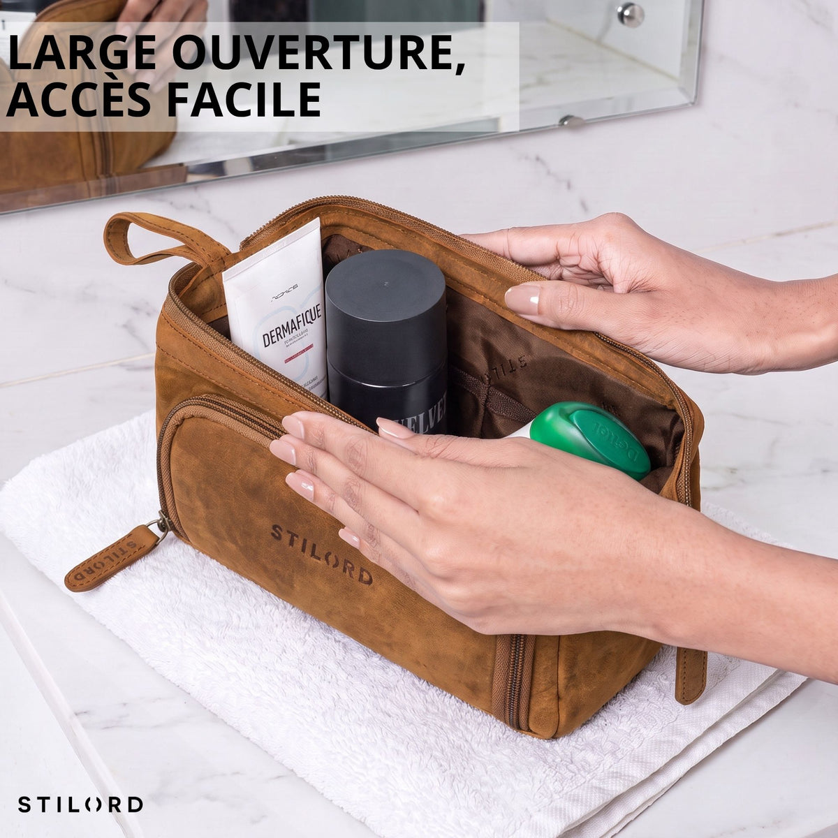 Trousse de toilette en cuir tan ouverte sur une serviette, mains écartent la fermeture, produits de soin visibles, marquage STILORD